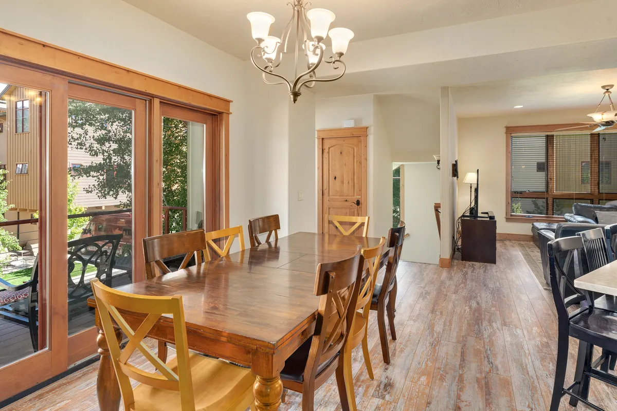 A bright dining area with a wooden table, plush chairs, a statement chandelier, and large sliding glass doors leading out to the deck. The setup encourages socializing and offers a direct visual cue to the adjoining deck, connecting indoor and outdoor living.