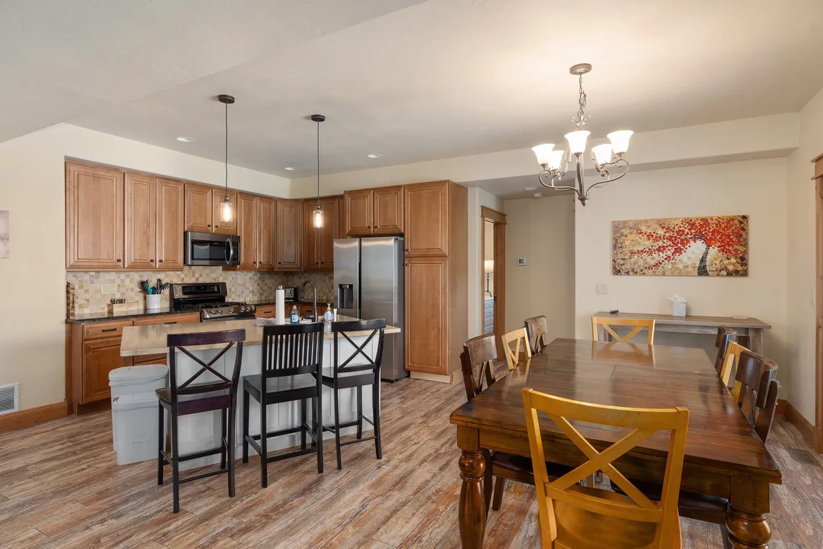 A wide-angle view of the open-concept main living space, showcasing the kitchen island, dining table with a striking chandelier overhead, stainless steel appliances, and tasteful artwork on the wall. The layout emphasizes the seamless connection between the kitchen and dining area, perfect for gatherings and shared meals.
