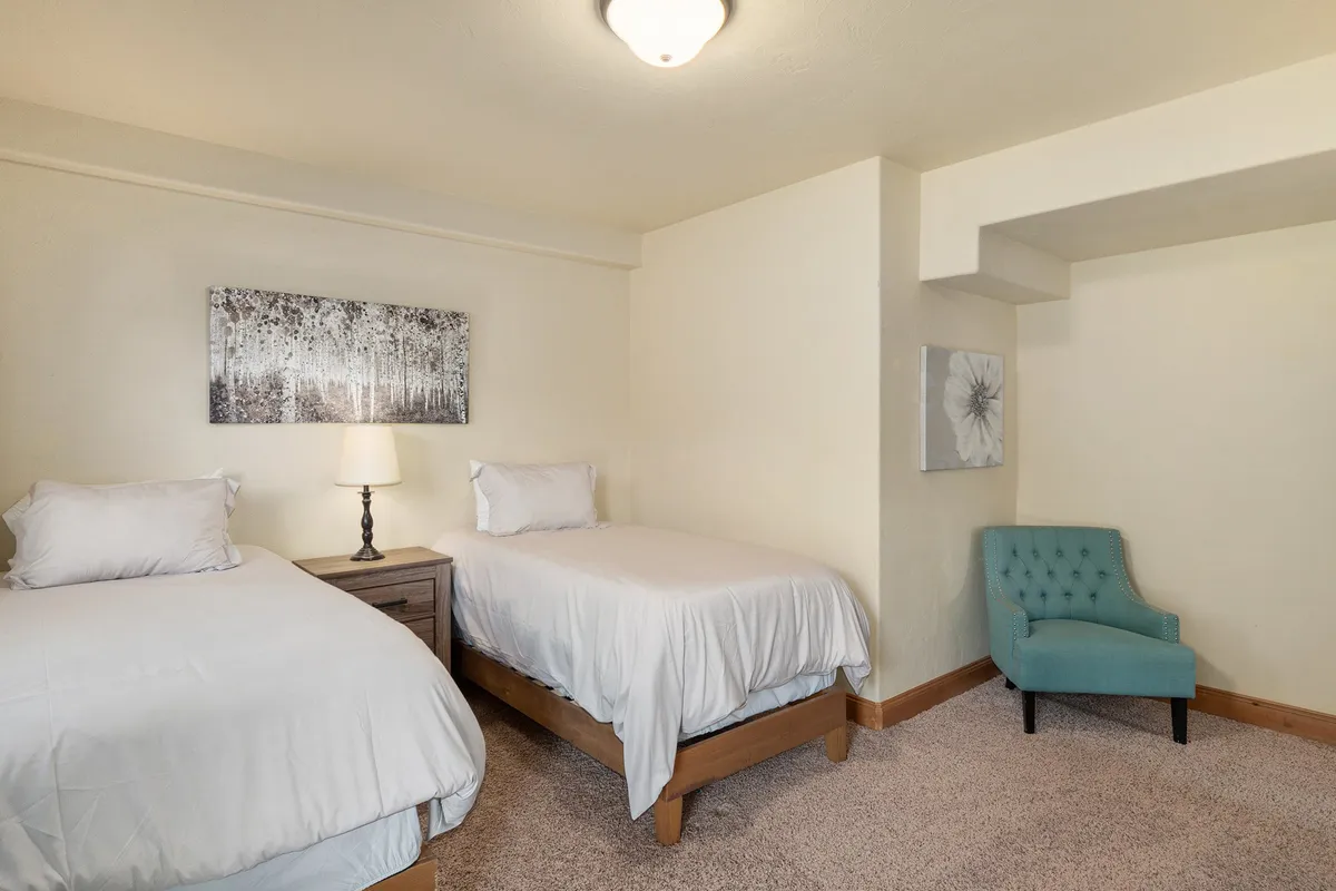 A serene twin suite featuring two twin beds, a stylish teal accent chair, a nightstand with a lamp, and thoughtful wall art. The atmosphere is quiet and comfortable, making it ideal for both restful nights and peaceful daytime reading.