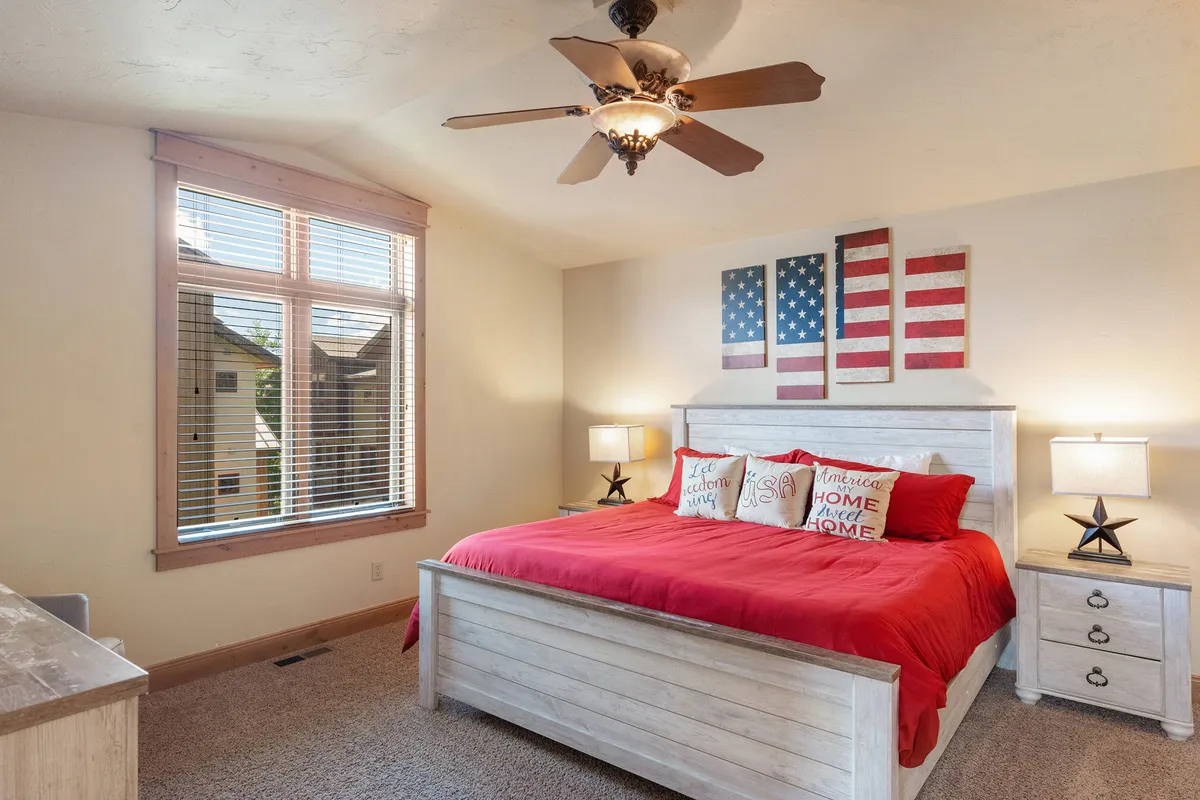 This image showcases a cozy bedroom with a red bedspread, flag-themed wall art, wooden furniture, and a ceiling fan. The room is filled with natural light, suggesting a peaceful space for relaxation or quiet work moments.